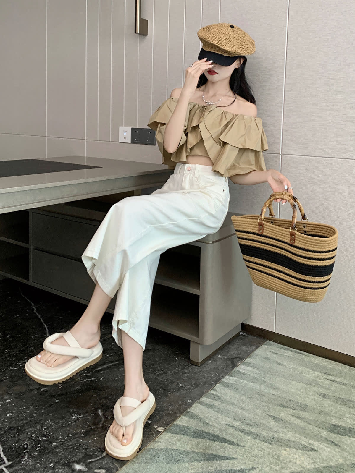 YEEYEEWang Wide-Leg Jeans in Ivory, Asian-fit cut, full-body seated pose on a desk edge, one hand at cap brim, striped straw tote in hand.