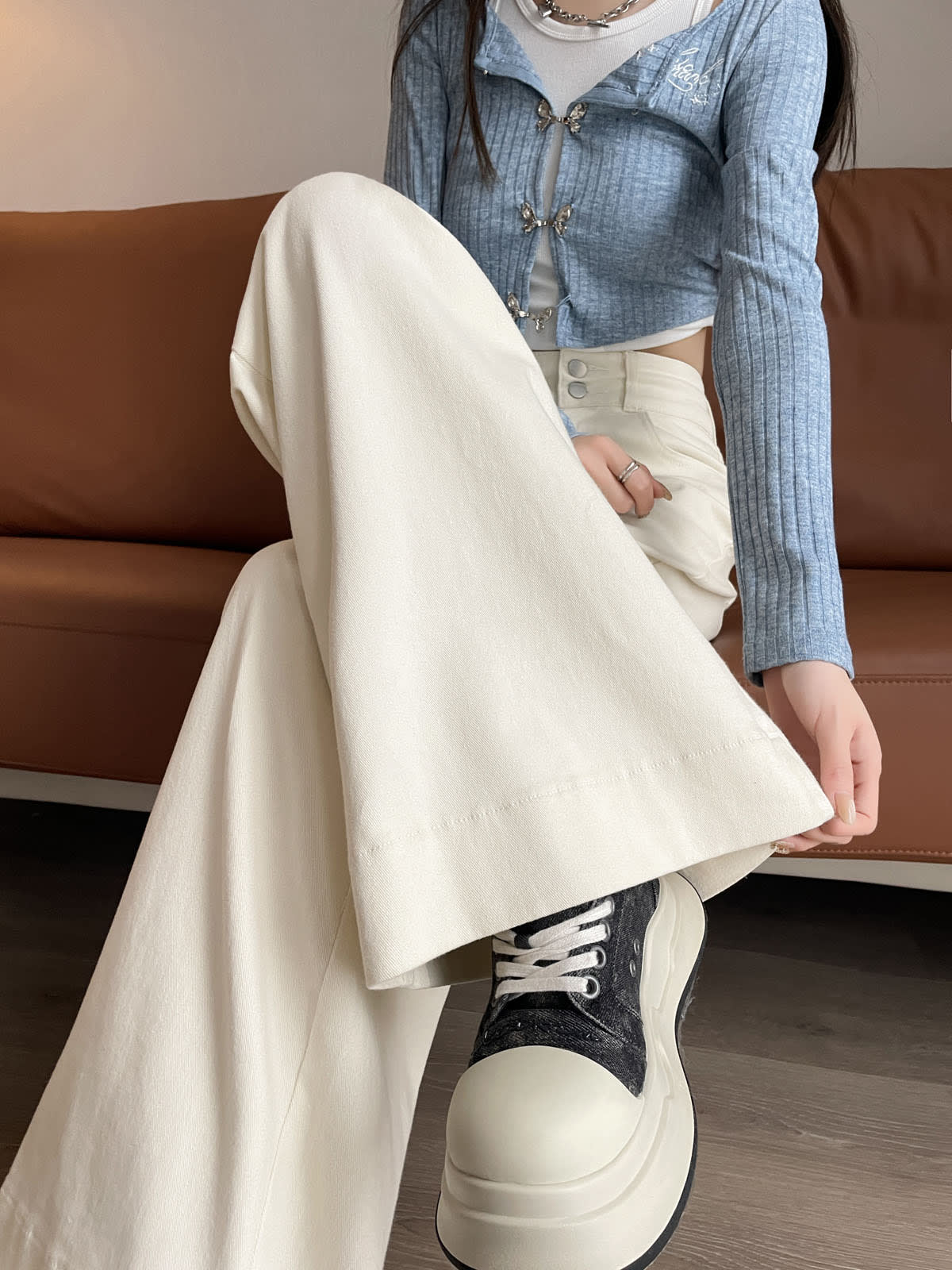 YEEYEEWang wide-leg jeans in Ivory, Asian-fit cut, waist-down seated pose on sofa, holding cuffed hem to show wide leg over chunky sneakers.