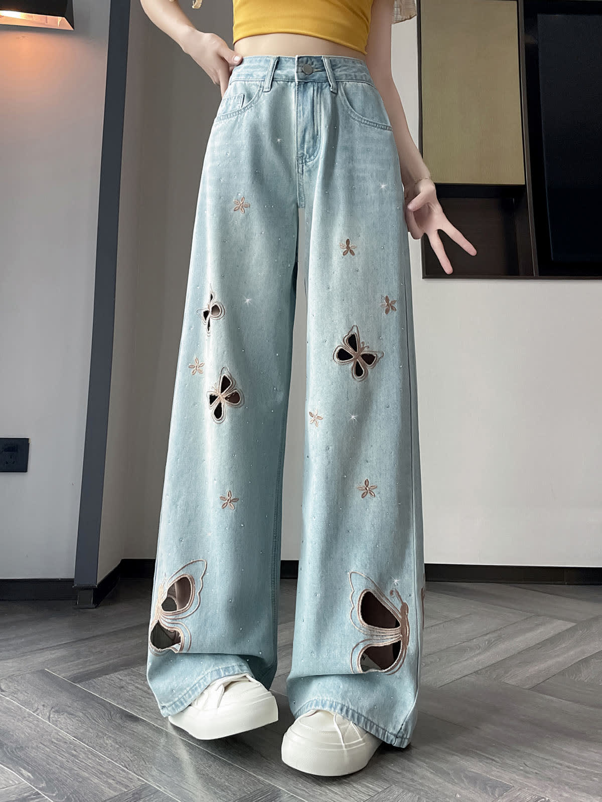 YEEYEEWang Wide-Leg Jeans in Light Blue, Asian-fit cut, waist-down front view with hand resting on waistband, butterfly cutout embroidery and scattered floral stitching, silver-tone button, floor-sweeping drape over white sneakers, variant thumbnail.