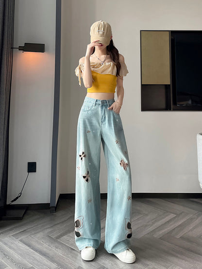YEEYEEWang Wide-Leg Jeans in Light Blue, Asian-fit cut, full-body standing pose with one hand in pocket and one touching cap brim, yellow off-shoulder ruffle top, butterfly cutouts and floral stitching down the legs.