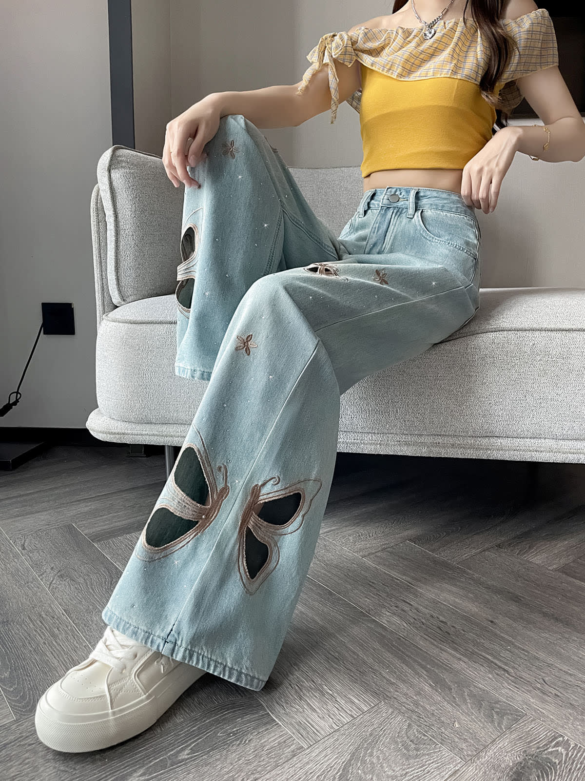 YEEYEEWang Wide-Leg Jeans in Light Blue, Asian-fit cut, waist-down seated pose on a gray sofa with legs extended forward, butterfly cutout embroidery on both legs, silver-tone button, white platform sneakers.