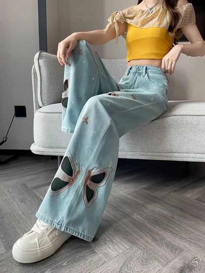 YEEYEEWang Wide-Leg Jeans in Light Blue, Asian-fit cut, waist-down seated pose on a gray sofa with legs extended forward, butterfly cutout embroidery on both legs, silver-tone button, white platform sneakers.