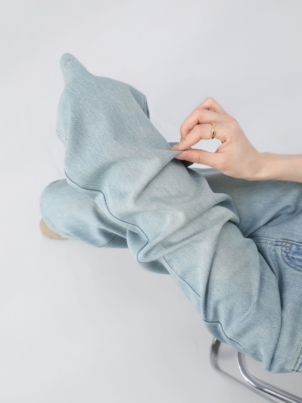 YEEYEEWang Wide-Leg Jeans in Light Blue, Asian-fit cut, close-up of hand pinching soft denim fabric at the knee to show texture, seated on chrome stool.