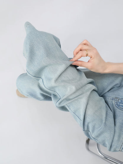 YEEYEEWang Wide-Leg Jeans in Light Blue, Asian-fit cut, close-up of hand pinching soft denim fabric at the knee to show texture, seated on chrome stool.