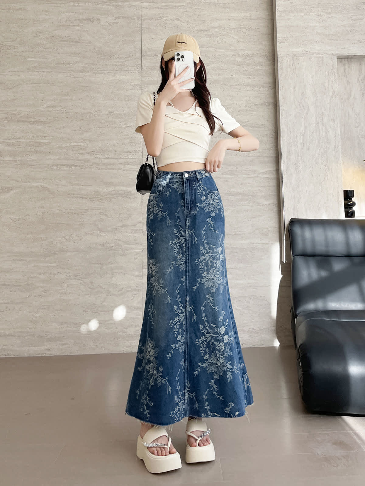 YEEYEEWang Jupe longue en jean Bleu, coupe Asian-fit, vue de face corps entier, debout avec téléphone devant le visage, main posée sur la ceinture, casquette beige et sac à chaîne noir, imprimé floral.