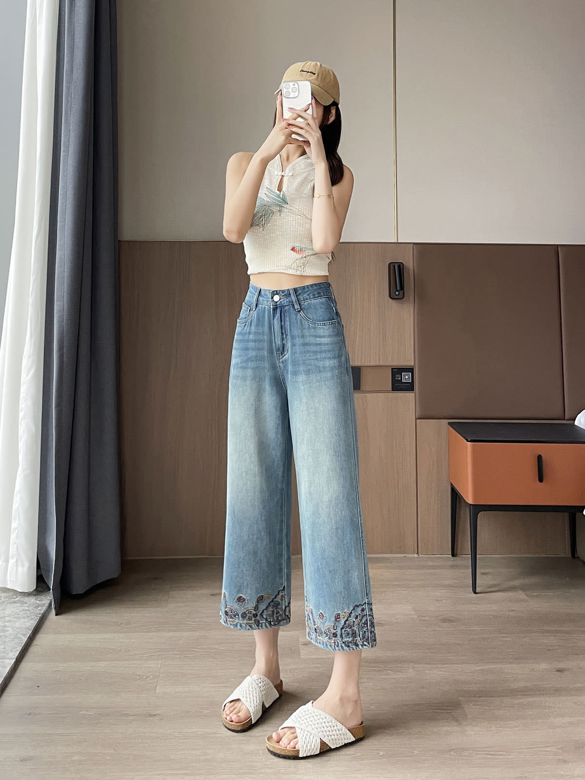 YEEYEEWang Straight Jeans in Vintage Blue, Asian-fit cut, full-body front standing pose holding phone at face with both hands, cap on head, floral-embroidered hems, indoor setting.