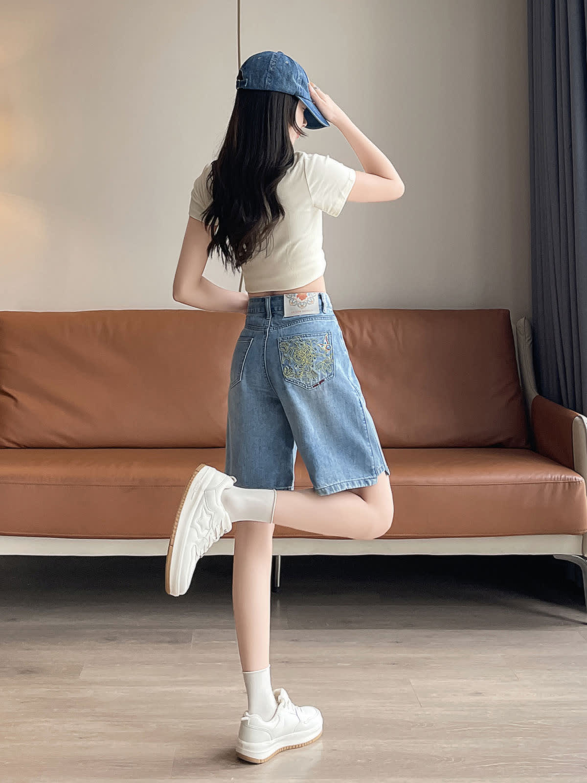 YEEYEEWang Denim Bermuda Shorts in Blue, Asian-fit cut, full-body back three-quarter leg-kick pose, hand touching the cap brim, embroidered back patch pocket visible.