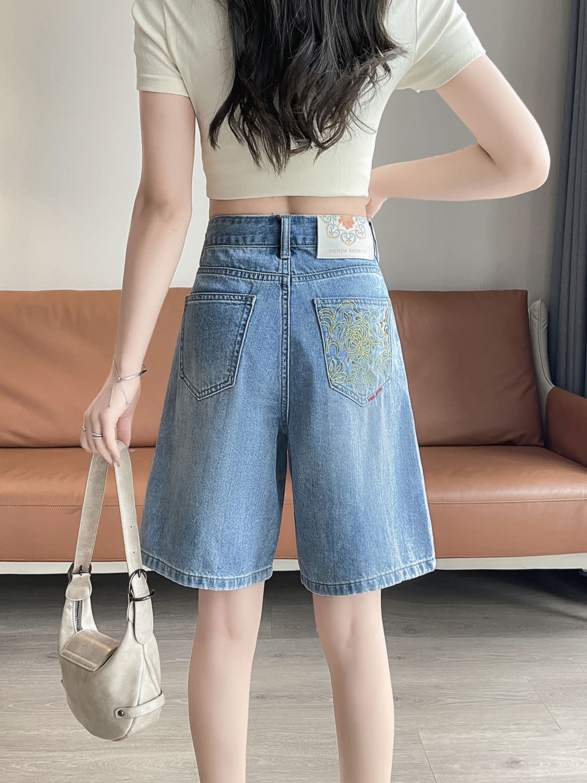 YEEYEEWang Denim Bermuda Shorts in Blue, Asian-fit cut, waist-down back view holding a cream handbag, embroidered back patch pocket and waistband label visible.