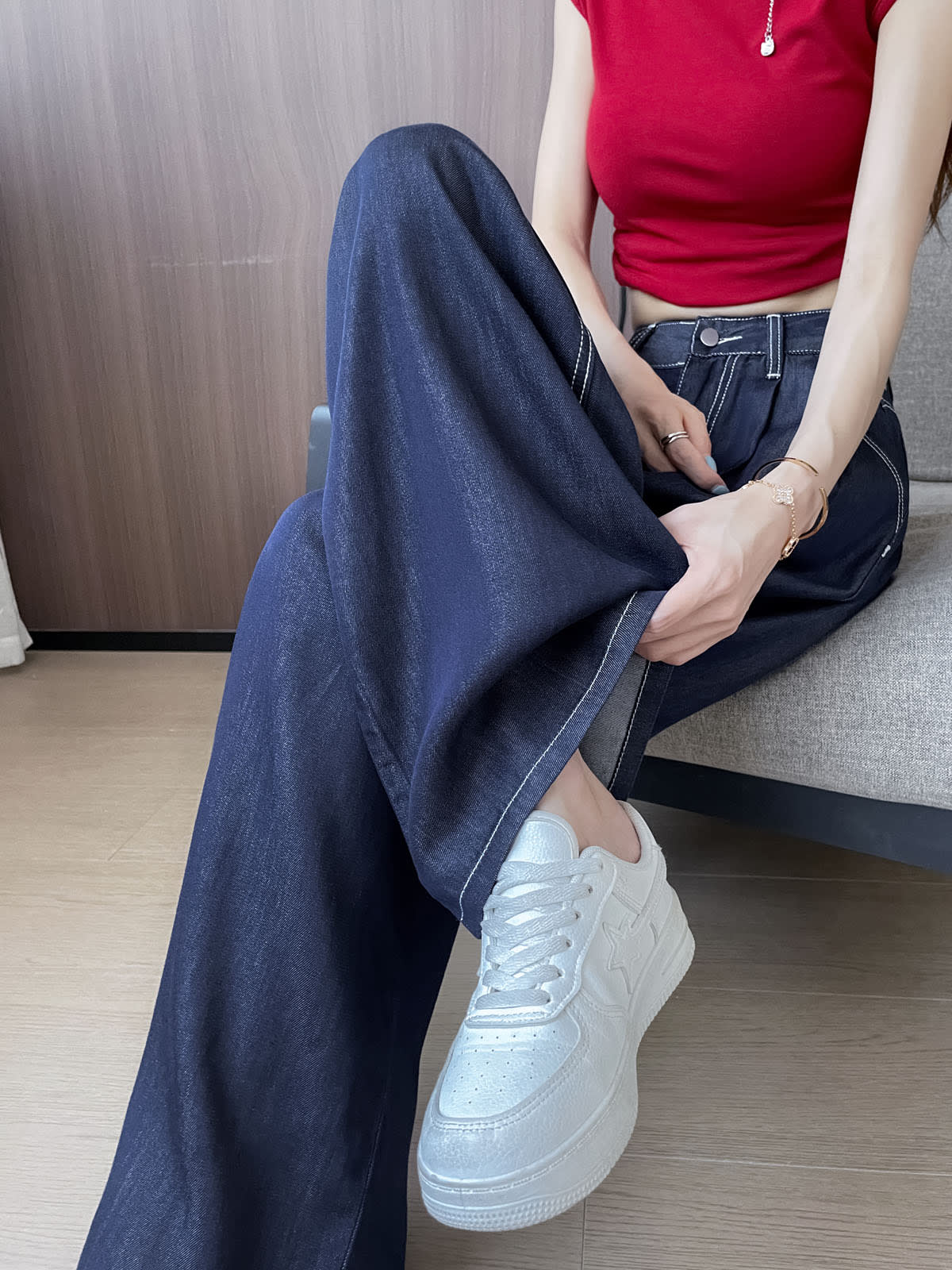 YEEYEEWang Wide-Leg Jeans in Denim Blue, Asian-fit cut, waist-down seated pose on sofa, one hand lifting the hem, paired with white sneakers.