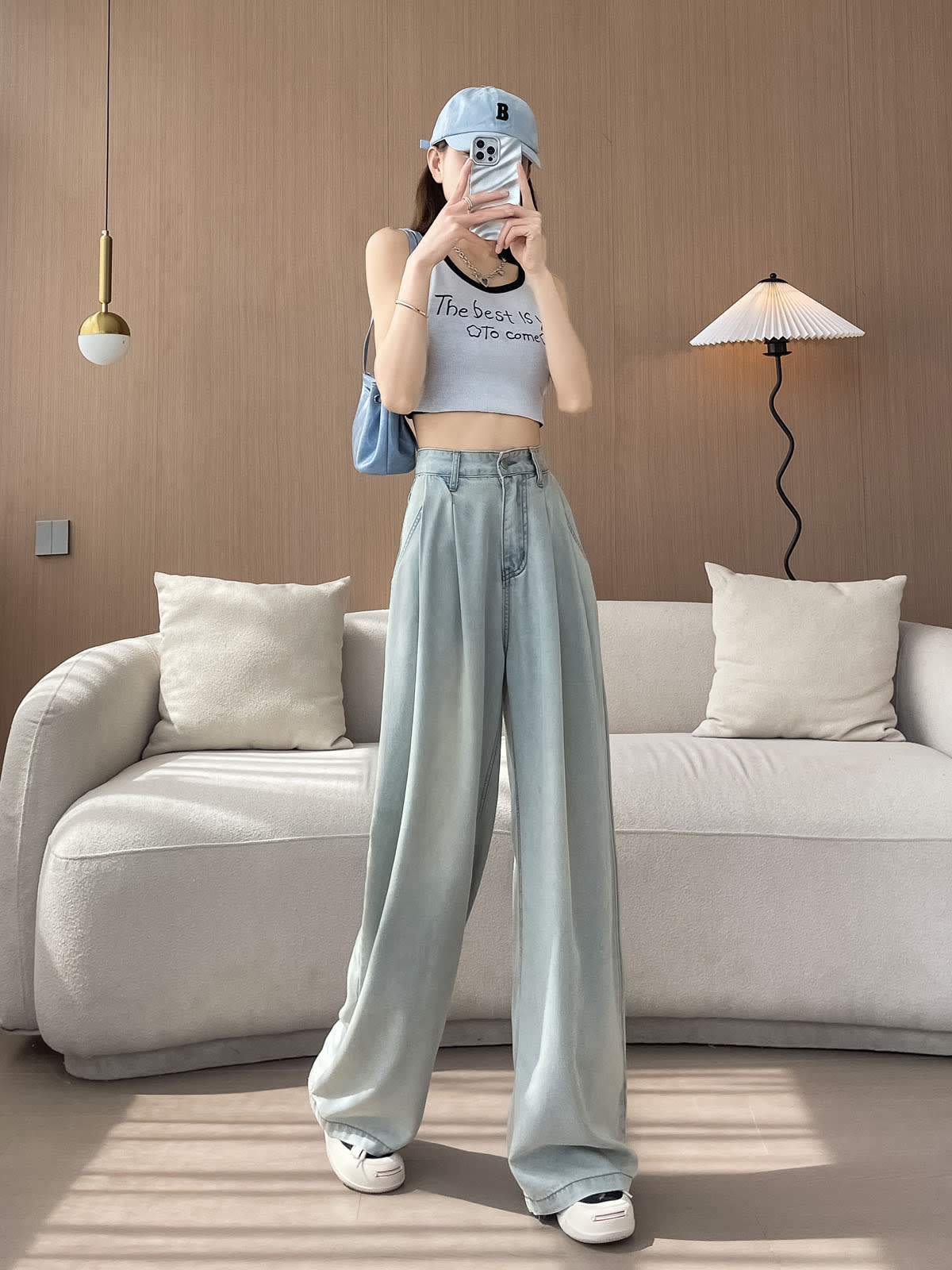 YEEYEEWang Wide-Leg Jeans in Light Blue, Asian-fit cut, full-body front pose with legs crossed holding phone at face, cap and blue bag styling.